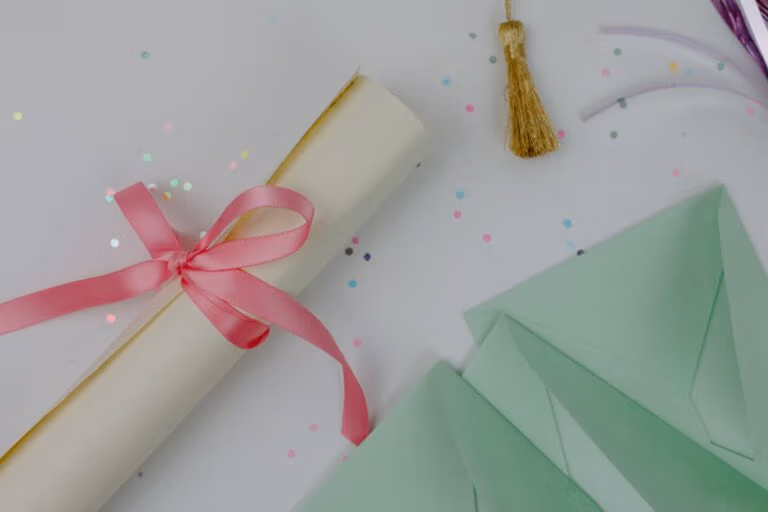 Flat lay of a decorative diploma with a pink ribbon, surrounded by confetti and green envelopes.